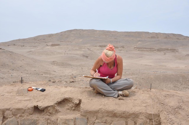 Estelle is sitting on the edge of an archaeological excavation in the desert writing on a clipboard, to her right there are some tools - a soft brush and tap measure. 