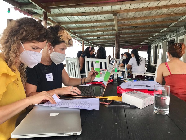 Two women are sat at a desk with laptops and face masks, behind them is a room full of people as part of a Sustainable Development Goals workshop in Galapagos